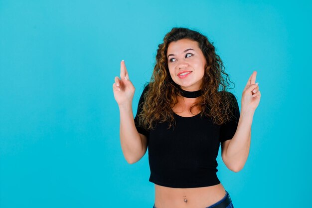 Lächelndes Mädchen schaut nach links, indem es die gekreuzten Finger auf blauem Hintergrund erhebt