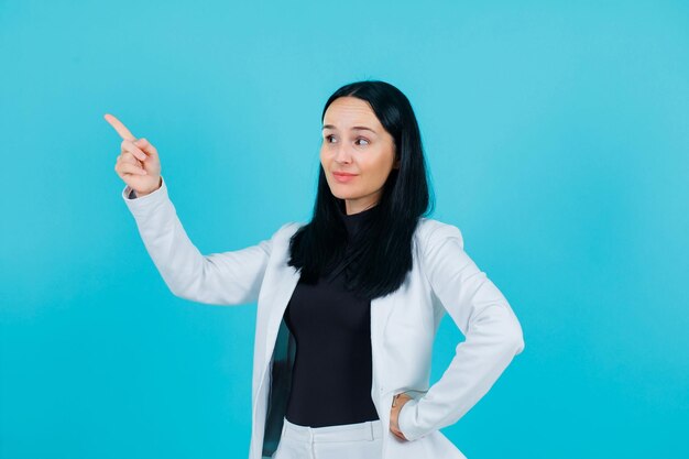 Lächelndes Mädchen zeigt mit dem Zeigefinger nach links und legt die andere Hand auf die Taille auf blauem Hintergrund