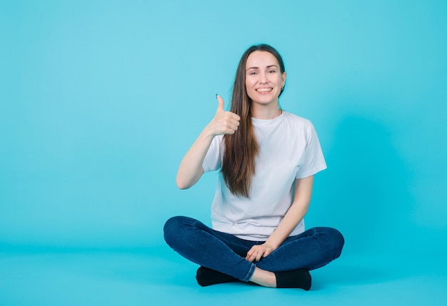 Lächelndes Mädchen zeigt perfekte Geste, indem es auf blauem Hintergrund auf dem Boden sitzt