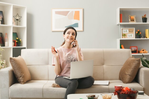 Lächelndes nachschlagendes junges Mädchen mit Laptop spricht am Telefon, das auf dem Sofa hinter dem Couchtisch im Wohnzimmer sitzt