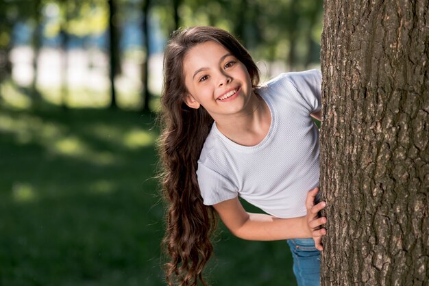 Lächelndes nettes Mädchen, das heraus von hinten Baumstamm draußen betrachtet