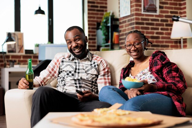 Lächelndes Paar, das sich Filme im Fernsehen ansieht und Essen zum Mitnehmen von der Lieferung isst und gemeinsam Spaß hat. Fast Food zum Mitnehmen und Bierflaschen genießen, während sie sich einen Film im Fernsehen ansehen.