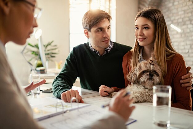 Lächelndes Paar mit einem Hund, das während eines Meetings im Büro mit seinem Versicherungsagenten spricht