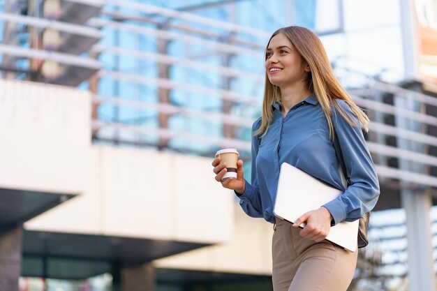 Lächelndes Porträt der blonden jungen Frau, das Laptop und Kaffee hält und blaues sanftes Hemd über modernem Gebäude trägt