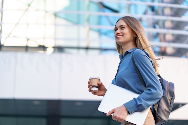 Lächelndes Porträt der blonden jungen Frau, das Laptop und Kaffee hält und blaues sanftes Hemd über modernem Gebäude trägt