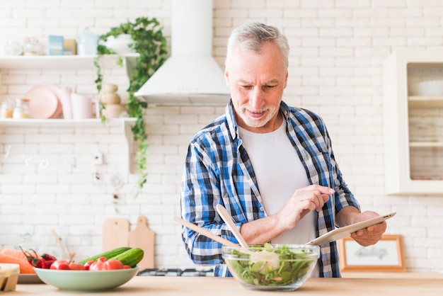 Lächelndes Porträt des älteren Mannes Lebensmittel unter Verwendung der digitalen Tablette in der Küche zubereitend