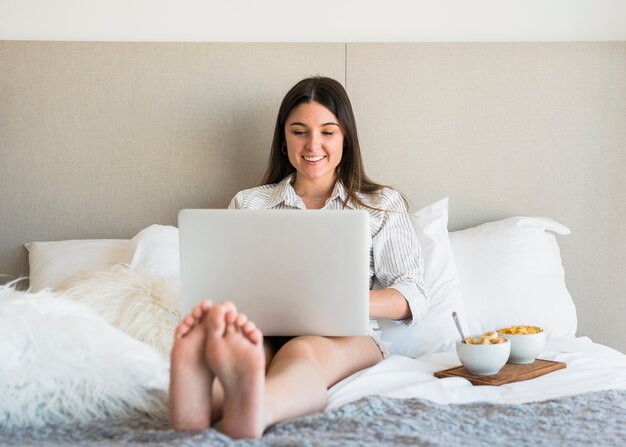 Lächelndes Porträt einer Frau, die auf Bett mit gesundem Frühstück unter Verwendung des Laptops sitzt