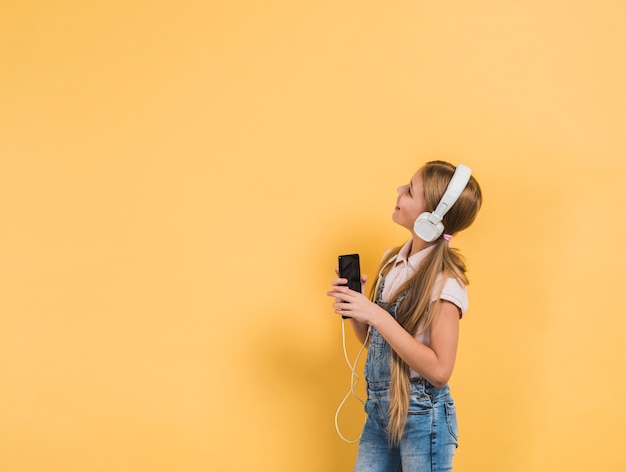 Lächelndes Porträt einer hörenden Musik des Mädchens auf dem Kopfhörer, der in der Hand Smartphone hält, gelben Hintergrund betrachtend