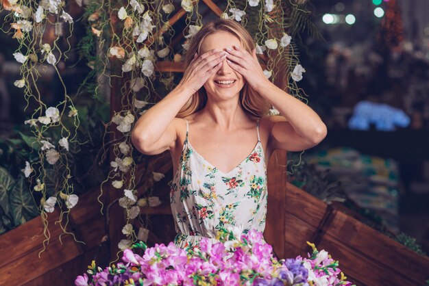 Lächelndes Porträt einer jungen Frau, die ihre Augen mit zwei Händen stehen vor Blumen bedeckt