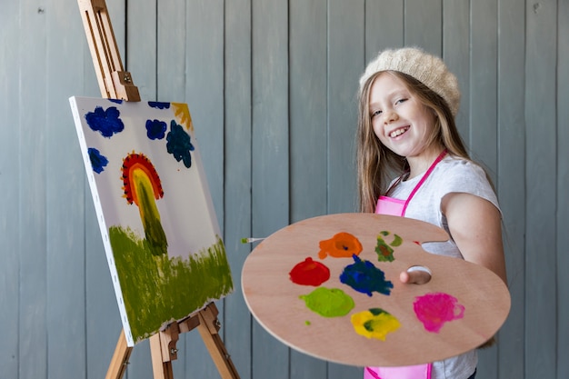 Lächelndes Porträt eines blonden Mädchens, das in der Hand die hölzerne Palette malt das Segeltuch hält