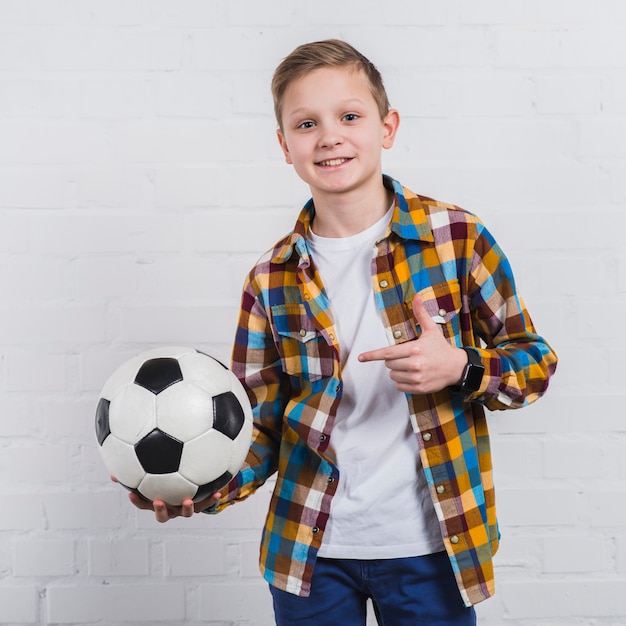 Lächelndes Porträt eines Jungen, der seinen Fußball steht gegen weiße Backsteinmauer zeigt