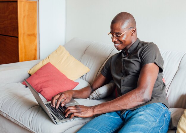 Lächelndes Porträt eines jungen Mannes, der auf Sofa unter Verwendung des Laptops sitzt