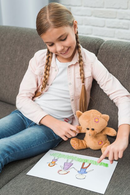 Lächelndes Porträt eines Mädchens, das auf dem Sofa zeigt die Familienzeichnung zum Teddybären sitzt