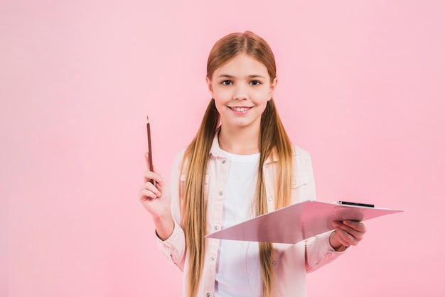 Lächelndes Porträt eines Mädchens, das Bleistift und Klemmbrett in den Händen gegen rosa Hintergrund hält