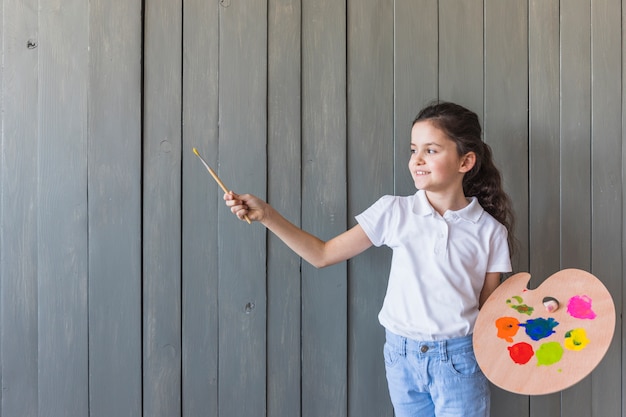 Lächelndes Porträt eines Mädchens, das den Pinsel und die Palette in der Hand stehen gegen graue hölzerne Wand hält