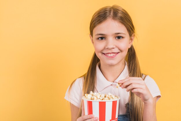 Lächelndes Porträt eines Mädchens, das Popcorneimer gegen gelben Hintergrund hält