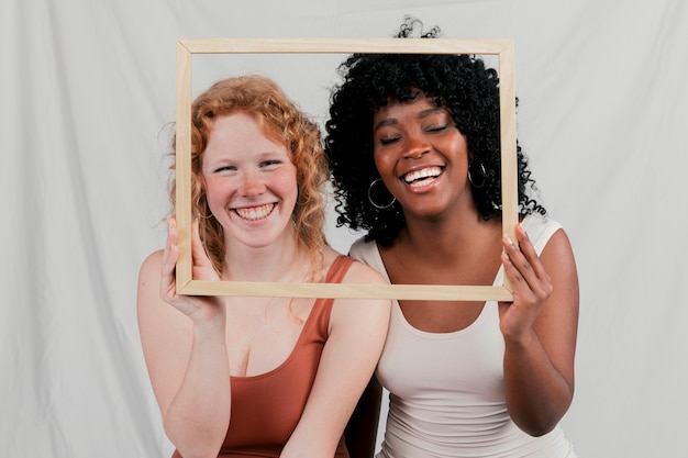 Lächelndes Porträt von den blonden und afrikanischen jungen Frauen, die Holzrahmen vor ihrem Gesicht halten