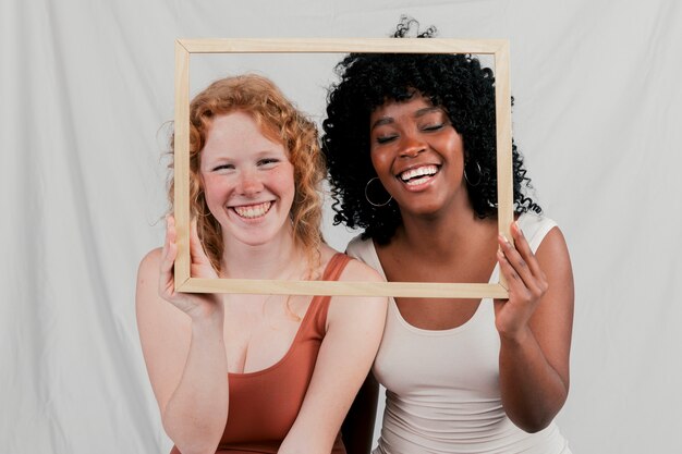 Lächelndes Porträt von den blonden und afrikanischen jungen Frauen, die Holzrahmen vor ihrem Gesicht halten