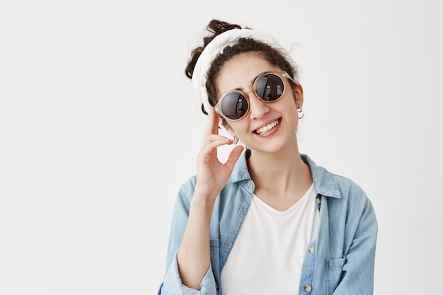 Lächelndes positives weibliches Model in trendiger runder Sonnenbrille mit Lappen im Jeanshemd, hat gute Laune, zeigt weiße Zähne, freut sich, Freunde und Verwandte zu treffen. Glück, Gesichtsausdruck Konzept