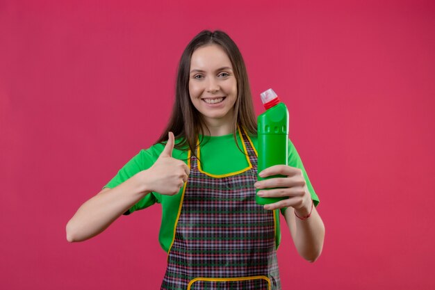 Lächelndes putzendes junges Mädchen, das Uniform hält, das Reinigungsmittel ihren Daumen oben auf lokalisiertem rosa Hintergrund hält