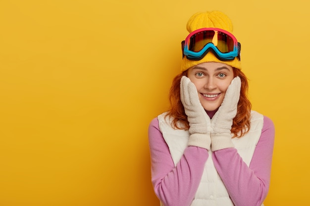 Lächelndes rothaariges Snowboardermädchen hat glücklichen Ausdruck, berührt Wangen, lokalisiert über gelbem Hintergrund