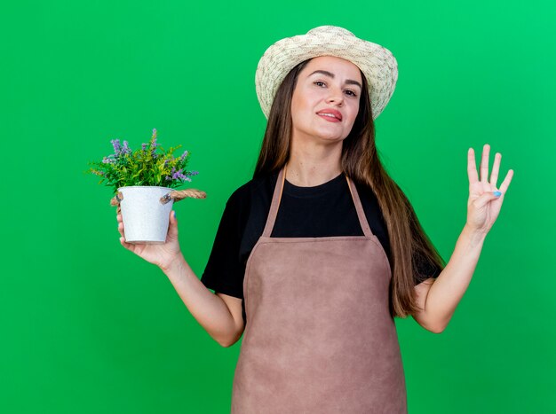 Lächelndes schönes Gärtnermädchen in der Uniform, die Gartenhut hält, der Blume im Blumentopf hält, der vier lokalisiert auf grünem Hintergrund zeigt