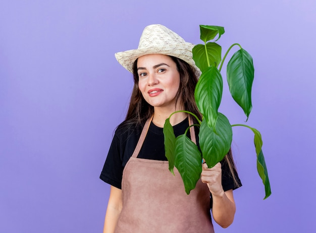 Lächelndes schönes Gärtnermädchen in der Uniform, die Gartenhuthaltepflanze lokalisiert auf Blau trägt