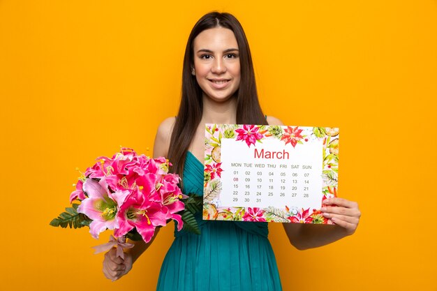 Lächelndes schönes junges Mädchen am Tag der glücklichen Frau, das Kalender mit Blumenstrauß lokalisiert auf orange Wand hält