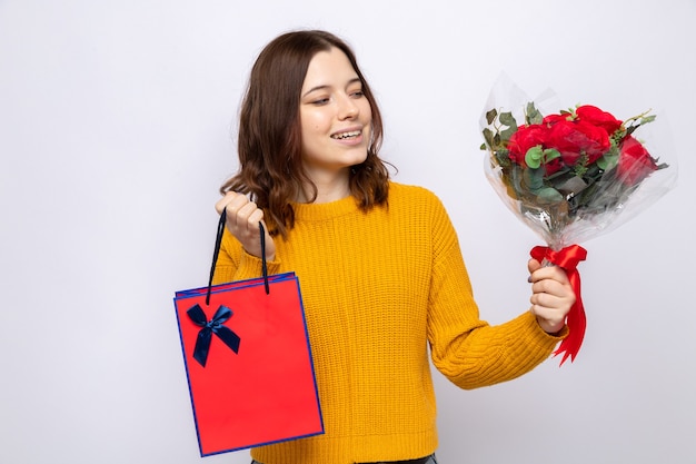 Lächelndes schönes junges Mädchen, das Geschenktüte hält und Blumenstrauß in der Hand betrachtet