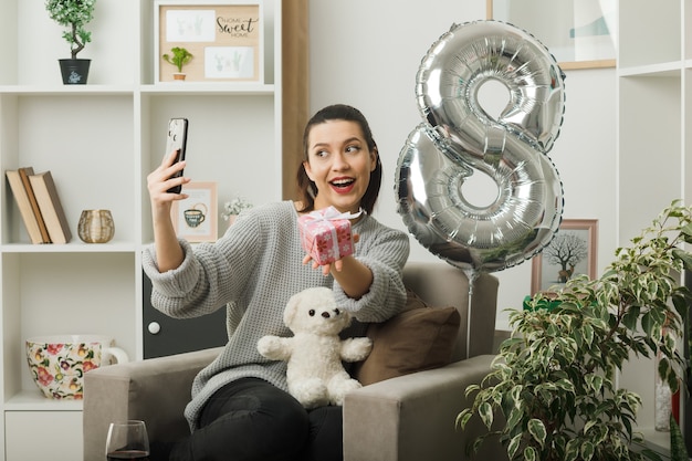 Lächelndes schönes Mädchen am glücklichen Frauentag mit Geschenk machen ein Selfie, das auf einem Sessel im Wohnzimmer sitzt