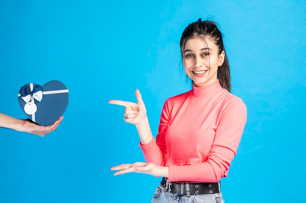 Lächelndes schönes Mädchen, das mit dem Finger auf die Geschenkbox auf blauem Hintergrund zeigt