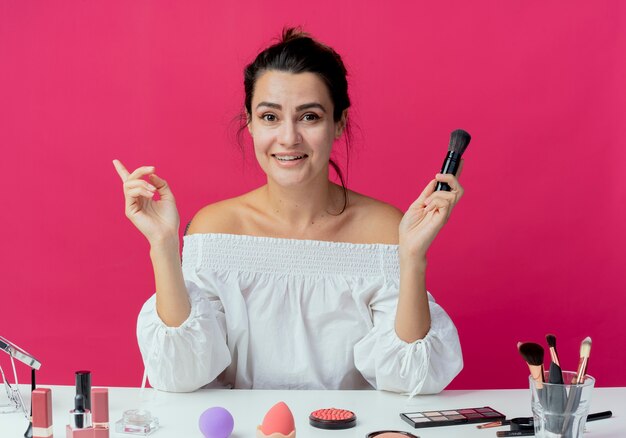 Lächelndes schönes Mädchen sitzt am Tisch mit Make-up-Werkzeugen, die Make-up-Pinsel halten und zur Seite zeigen, die auf rosa Wand lokalisiert wird