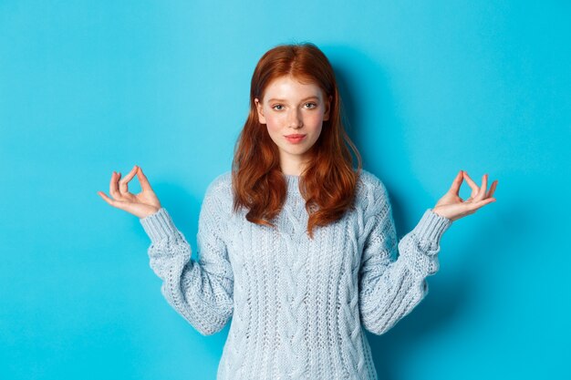 Lächelndes selbstbewusstes Mädchen mit roten Haaren, das geduldig bleibt, Händchen in Zen hält, Meditationspose und in die Kamera starrt, Yoga praktiziert und ruhig vor blauem Hintergrund steht.