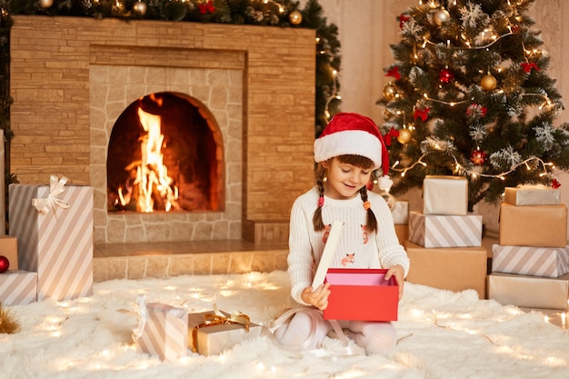 Lächelndes süßes kleines Mädchen mit weißem Pullover und Weihnachtsmann-Hut, das im festlichen Raum mit Kamin und Weihnachtsbaum posiert und geöffnete Weihnachtsgeschenkbox hält.