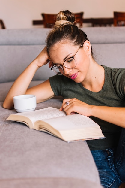 Lächelndes tragendes Brillenlesebuch der jungen Frau nahe dem Sofa