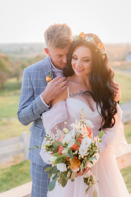 Lächelndes zartes Hochzeitspaar verliebt im Freien auf der Wiese mit schönem Hochzeitsstrauß und Kranz am sonnigen Tag