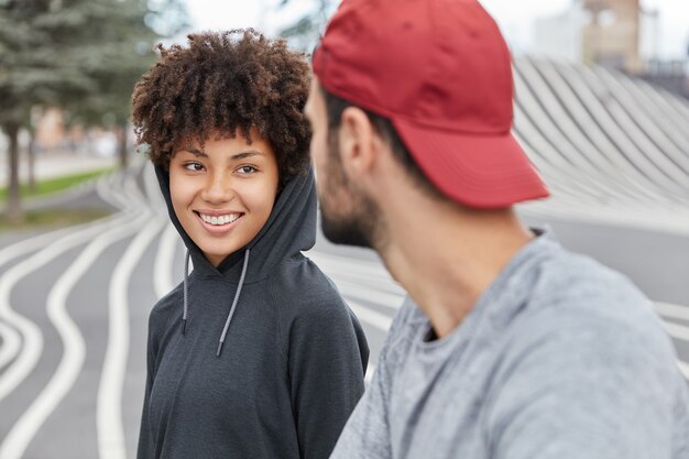 Lächelndes zufriedenes ethnisches süßes Mädchen im Kapuzenpulli hat angenehmes freundliches Gespräch mit einem Kerl, gehen Sie im Freien