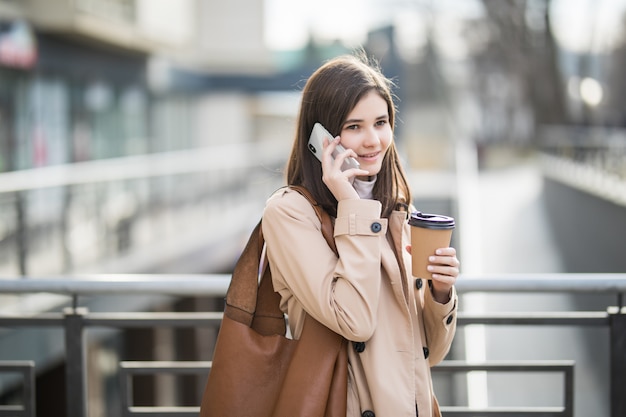 Lässig gekleidete Frau, die die Straße geht, die Kaffeetasse und Telefon hält