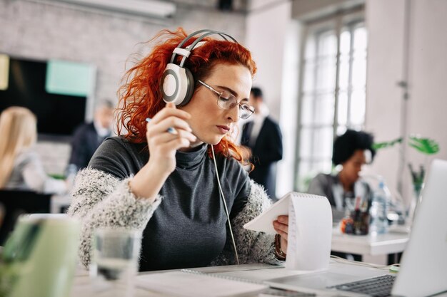 Lässige Geschäftsfrau, die an ihrem Schreibtisch arbeitet und Notizen liest, während sie Musik über Kopfhörer hört. Es gibt Menschen im Hintergrund