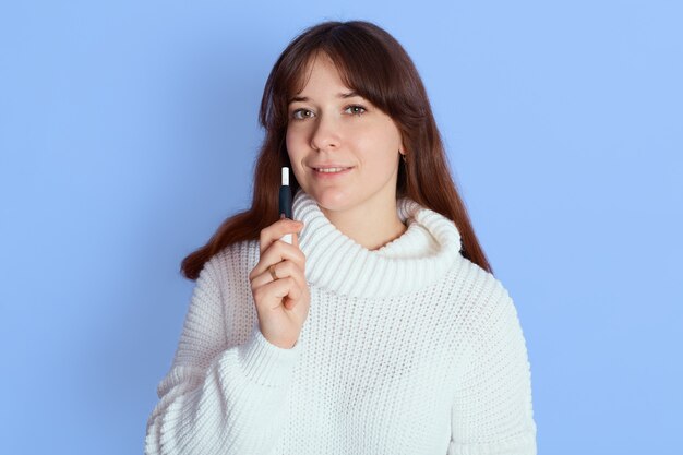 Lässige hübsche Frau, die steht und über Blau vaping, während sie direkt in die Kamera schaut, Mädchen mit dunklem Haar kleidet weißen Pullover, hält e Zigarette.