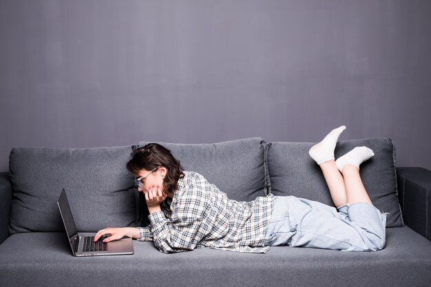 Lässige junge Frau barfuß, die auf ihrem Bauch liegt, der auf einem Sofa unter Verwendung ihres Laptops entspannt