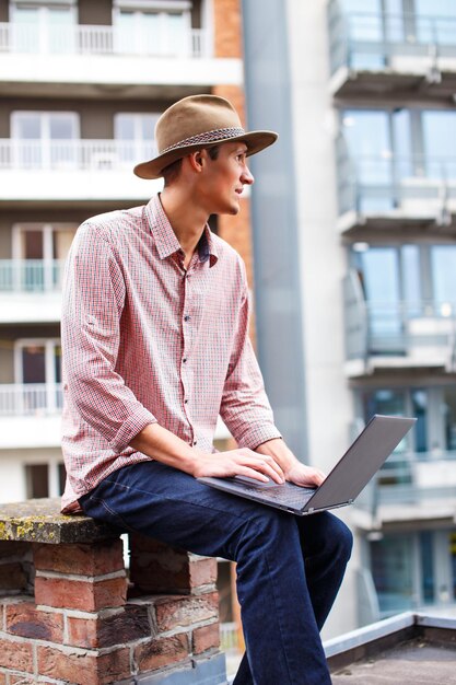 Lässiger Mann mit Hut, der einen Laptop auf dem Dach gegen ein Stadtgebäude hält.