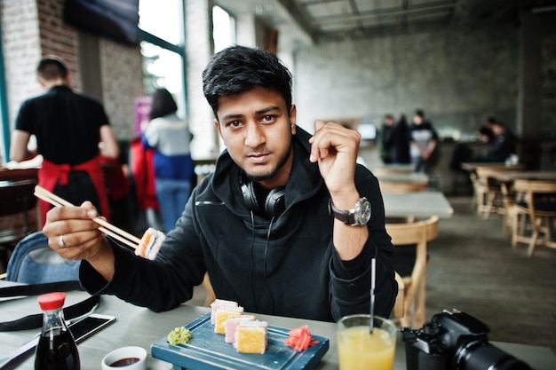 Lässiger und stilvoller junger asiatischer Mann mit Kopfhörern im Café, das Sushi isst
