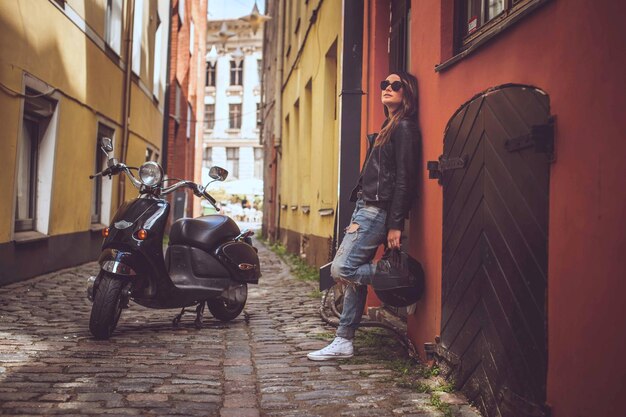 Lässiges Mädchen mit Sonnenbrille, das einen schwarzen Moto-Helm hält und mit einem Moto-Roller in der Nähe einer alten roten Wand posiert.