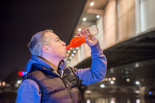 Kostenloses Foto läufer in blau sportkleidung trinken wasser und stehen in der nähe des flusses. hübscher senior jogging mann trinkt frisches wasser aus flasche nach dem laufen