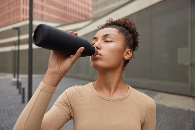 Läuferin mit lockigem gekämmtem Haar fühlt sich nach intensivem Training durstig und trinkt frisches Wasser aus der Flasche, die in Sportbekleidung gekleidet ist und im Freien vor verschwommenem Hintergrund posiert Sportlerin nach dem Training