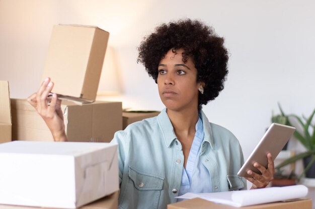 Lageristin mit Afrofrisur-Prüfpaket. Ernsthafte junge Frau, die ein Tablet verwendet, während sie zwischen Stapeln von Kisten arbeitet. Lagerkonzept