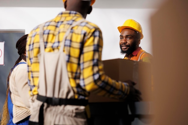 Lagerleiter sieht Kollegen an, die Pakete zur Rezeption im Lagerraum bringen. Afroamerikanischer Lagerhausangestellter mit schützendem Schutzhelm, der eine Box vom Pakethändler erhält