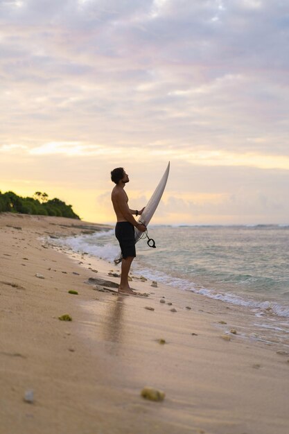 Landschaftsbild eines männlichen Surfers, der bei Sonnenaufgang am Strand spazieren geht, während er sein Surfbrett unter dem Arm trägt, während die Meereswellen im Hintergrund brechen. Junger hübscher männlicher Surfer auf dem Ozean