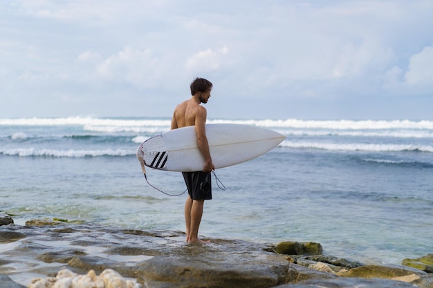 Landschaftsbild eines männlichen Surfers, der bei Sonnenaufgang am Strand spazieren geht, während er sein Surfbrett unter dem Arm trägt, während die Meereswellen im Hintergrund brechen. Junger hübscher männlicher Surfer auf dem Ozean